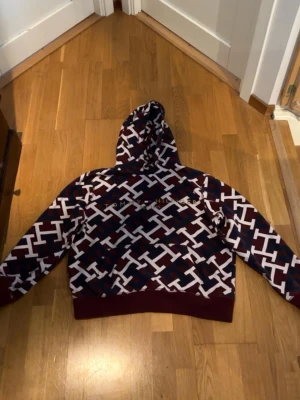 Mönstrad hoodie från Tommy Hilfiger - Säljer en snygg hoodie från Tommy Hilfiger i storlek M. Hoodien har ett coolt mönster i vinrött, marinblått och vitt med stora loggor över hela plagget. Den har huva med dragsko och breda muddar. Perfekt för dig som vill sticka ut lite extra.