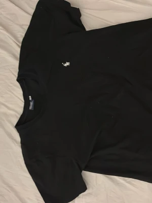 Svart t-shirt från Polo Ralph Lauren - Klassisk svart t-shirt från Polo Ralph Lauren med den ikoniska vita broderade loggan på bröstet. T-shirten har rund hals och korta ärmar, perfekt för en clean och stilren look. Tillverkad i mjuk bomull för maximal komfort. Storleken är i M men passar S också 
