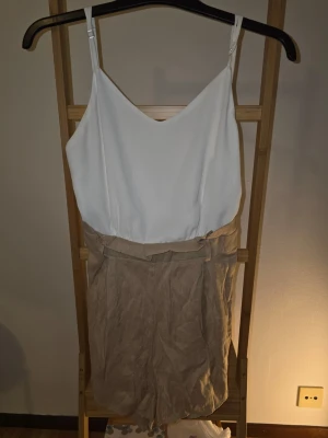 Beige och vit jumpsuit med smala band - Snygg jumpsuit med vit topp och beige shortsdel. Tunt tyg, smala justerbara axelband och v-ringad hals. Midjan har ett bälte i samma material som shortsen. Perfekt för en avslappnad och trendig look. Snöret till fattas.