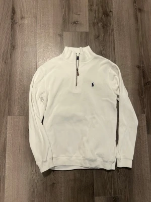 Vit half-zip tröja från Polo Ralph Lauren - Klassisk vit tröja med half-zip och hög krage från Polo Ralph Lauren. Tröjan har det ikoniska mörkblå broderade logot på bröstet och ribbade muddar vid ärmslut och nederkant. Perfekt för en clean och stilren look.