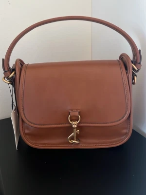 Brun handväska från Zara  - Snygg brun handväska från Zara i slätt läder med guldfärgade metalldetaljer och ett klassiskt spänne framtill. Väskan har ett stadigt handtag och stilren, boxig form. Perfekt accessoar för dig som gillar tidlös design och exklusiva material.