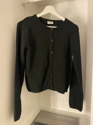 Svart kofta från Kappahl - En enkel och stilren svart kofta från Kappahl med rund halsringning och knäppning framtill. Koftan har långa ärmar och ribbade muddar vid hals och ärmslut. Perfekt att slänga över en t-shirt eller linne för en clean look.