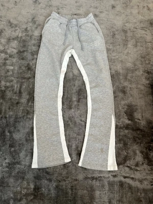 Flared Mjukisbyxor - Snygga grå mjukisbyxor med vita ränder längs benen. Byxorna har utsvängda ben och en liten broderad logga på vänster sida. Perfekta för en chill och avslappnad stil.