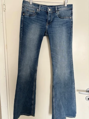 Blå bootcut jeans med låg midja - Säljer ett par klassiska blå bootcut jeans med låg midja . Jeansen har en snygg tvättad look och vida ben som ger en retrovibe. Perfekta för dig som vill ha en avslappnad men trendig stil. Materialet är mjukt denim i bomull.