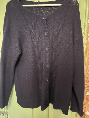 Mörklila kabelstickad kofta med knappar - Mysig mörklila kabelstickad kofta med långa ärmar och knäppning framtill. Koftan har ett klassiskt flätmönster längs knappraden och bred ribbad kant vid halsen. Väl använd men fortfarande fin. Lite sliten kring armbågarna. Be gärna om bilder! Storlek M. Priset kan alltid diskuteras!