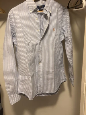 Blårandig skjorta från Polo Ralph Lauren - Klassisk blårandig skjorta från Polo Ralph Lauren med button-down krage och broderad logga på bröstet. Skjortan har lång ärm, vita knappar och är tillverkad i bomull. Perfekt till jeans eller chinos för en clean och stilren look.