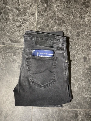 Grå jeans från Jacob Cohen - Snygga grå jeans från Jacob Cohen med klassisk femficksdesign och raka ben. Jeansen har en svart patch med logga bak i midjan och är tillverkade i mjukt denimtyg. Perfekta för dig som gillar stilrena och tidlösa plagg. Storleken är 31 och byxorna är i extremt bra skick! Bara att höra av sig vid eventuella frågor.
