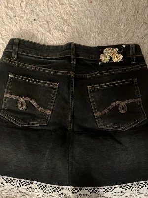 Svart jeanskjol med spets från Street One - Snygg svart jeanskjol från Street One med dekorativa bakfickor och vit spetskant längst ner. Kjolen har klassisk femficksdesign, bälteshällor och kontrastsömmar. Perfekt för dig som vill ha en cool och unik look.