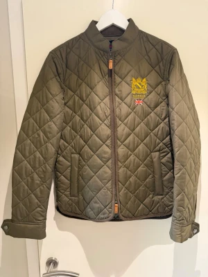 Olivgrön quiltad vindjacka Morris - Snygg olivgrön quiltad vindjacka från Morris med broderad logga och brittisk flagga på bröstet. Jackan har ståkrage, dragkedja framtill, två fickor och knappdetaljer vid ärmslut. Tillverkad i ett lätt och slitstarkt material, perfekt för lager-på-lager.
