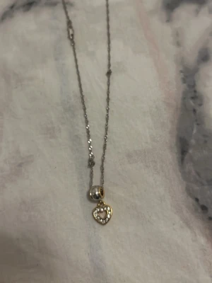 Silverhalsband med hjärthänge - Snyggt halsband i silverfärg med ett litet hjärthänge i guldton. Hänget är dekorerat med små gnistrande stenar runt kanten och har en öppen design. Kedjan är tunn och elegant, perfekt för att ge en söt touch till din outfit. Och även äkta rostfritt silver i mycket bra skick som jag fick i present 