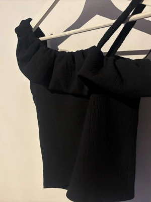 Svart oneshoulder topp med volang - Trendig svart oneshoulder topp med bred volang längs halsringningen och smal axelrem. Ribbad struktur i tyget ger en snygg detalj. Perfekt för dig som vill sticka ut med en cool och modern look.