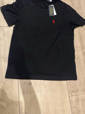 Svart t-shirt från Polo Ralph Lauren - Klassisk svart t-shirt från Polo Ralph Lauren med röd broderad logga på bröstet. T-shirten har rund halsringning och korta ärmar. Tillverkad i mjuk bomull för en skön känsla och enkel stil.
