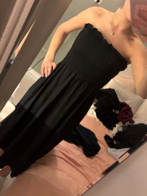 Svart  klänning - Säljer en svart axelbandslös klänning med smockad överdel och avtagbara axelband. Klänningen har en mjuk, flowy passform och är i midi-längd. Perfekt för sommarens alla tillfällen och enkel att styla med accessoarer.