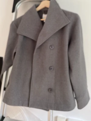 Grå kappa från Gina Tricot - Stilren grå kappa från Gina Tricot med bred krage och dubbelknäppning framtill. Klassisk rak modell i mjukt material som passar perfekt till höstens och vinterns outfits. Enkel att matcha och ger en snygg look.