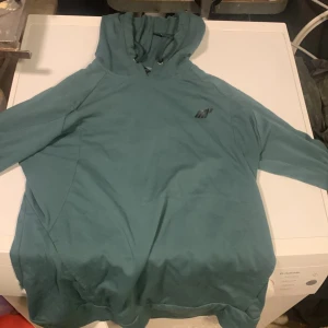 Blågrön hoodie från MyProtein - Snygg blågrön hoodie från MyProtein med diskret logga på bröstet. Hoodien har huva med dragsko och långa ärmar. Tillverkad i mjukt material som känns skönt mot huden. Gymtröja, träningskläder träningshoodi