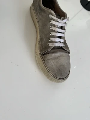 Gråa lanvin sneakers i mocka - Säljer ett par gråa sneakers i mocka med vita skosnören och vit sula. Klassisk låg modell med enkel design och diskreta sömmar. Perfekta för dig som gillar stilrena och neutrala skor. Tyvvärr rätt slitna med mycket skråmor. Därför priset är billigt och dom är ju perfekta för dig som vill fixa upp dom o sälja den dyrt! Pris ej hugget i sten