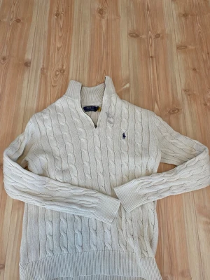 Vit kabelstickad tröja Polo Ralph Lauren - Kabelstickad vit tröja från Polo Ralph Lauren med halv dragkedja och hög krage. Tröjan har det klassiska broderade logotypen på bröstet och ribbade muddar vid ärmslut och nederkant. Perfekt för en stilren och avslappnad look.