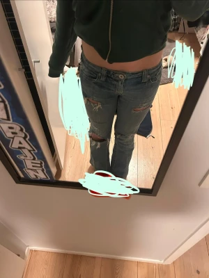 Blå slitna bootcut jeans - Säljer ett par vintage blå bootcut jeans med slitningar och hål på låren och knäna. Jeansen har låg midja och klassiska fickor både fram och bak, med snygga broderade detaljer på bakfickorna. Jeansen är köpta i USA och jag säljer dem eftersom de är lite för korta för mig.