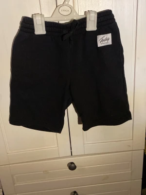 Svarta shorts från Andy by Frank Dandy - Svarta shorts med snörning i midjan från Andy by Frank Dandy. De har en enkel design med en liten vit logotyp på vänster sida och är tillverkade i mjukt material som passar perfekt för en avslappnad stil.