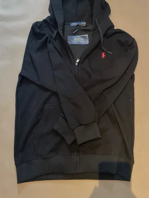 Svart hoodie med zip från Polo Ralph Lauren - Svart hoodie med dragkedja från Polo Ralph Lauren. Klassisk design med röd broderad logga på bröstet, huva med dragsko och kängurufickor. Tillverkad i mjuk bomull för skön känsla och stilren look. Helt oanvänd med tags kvar, pris kan diskuteras 