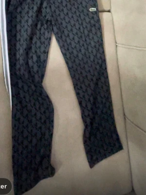 Svarta mjukisbyxor från Lacoste - Säljer ett par svarta mjukisbyxor från Lacoste med diskret mönster och vita sidoränder. Byxorna har en avslappnad passform och det klassiska Lacoste-logotypen på höften. Perfekta för chill dagar eller streetwear-stil.