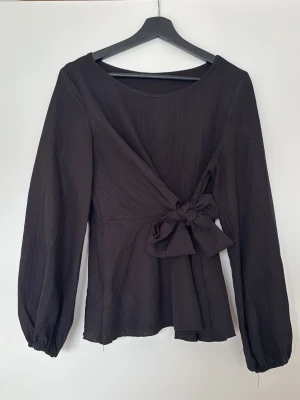 Svart omlottblus med knytband - Stilren svart omlottblus med långa ärmar och breda knytband i midjan. Blusen har en lätt struktur i tyget och rund halsringning. Finns även en dragkedja i ena sidan för enkel på och avtagning. Perfekt för dig som gillar en enkel men trendig look.