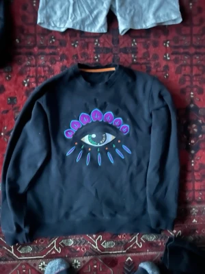 Svart sweatshirt |KENZO PARIS  - Cool svart sweatshirt med ett stort ögonmotiv i blått, lila och turkost på bröstet. Tröjan har rund halsringning och ribbade muddar vid ärmslut och nederkant. Perfekt för dig som gillar statement-plagg och grafiska tryck.