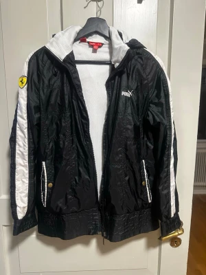 Svart Ferrari x Puma vindjacka - Svart vindjacka från Puma i samarbete med Ferrari. Jackan har vita detaljer längs ärmarna, Ferrari-logga på ärmen och ryggen samt en rand i Italiens färger på baksidan. Huva, dragkedja och fickor med tryckknappar. Materialet är glansigt och lätt. Den är ganska liten så skulle säga att den passar S-M. Armarna är lite smala för L