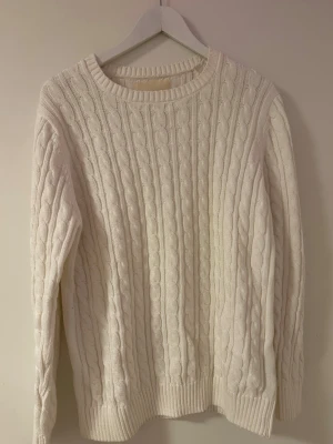 Vit kabelstickad tröja - Kabelstickad tröja i mjuk vit färg med rund halsringning och ribbade muddar vid ärmslut och nederkant. Klassisk och tidlös design som passar perfekt till jeans eller kjol. Materialet känns mjukt och bekvämt, perfekt för kyligare dagar.