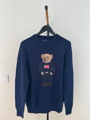 Mörkblå stickad tröja med björn Polo Ralph Laureb - Mörkblå stickad tröja från Polo Ralph Lauren med ikonisk björn på bröstet, klädd i blå outfit och amerikansk flagga på tröjan. Rund halsringning och ribbade muddar vid ärmslut och nederkant. Perfekt för dig som gillar klassiska och lekfulla detaljer.