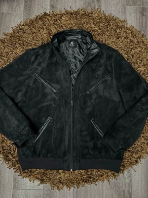 Svart skinnjacka från AC - Snygg svart skinnjacka från AC med fyra dragkedjeförsedda fickor framtill och ribbade muddar vid ärmslut och nederkant. Jackan har en hög krage och stilren design som passar till jeans eller byxor. Perfekt för dig som gillar en klassisk men modern look.