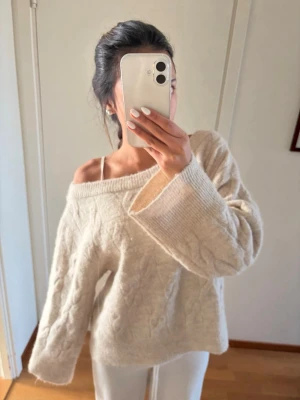 Beige kabelstickad oversized tröja - Mysig beige kabelstickad tröja med bred ribbad kant i halsen och vid ärmsluten. Tröjan har en avslappnad, oversized passform och kan bäras off-shoulder för en trendig look. Perfekt för kalla dagar och enkel att styla med olika outfits.