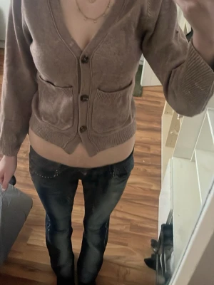 Brun kofta med knappar och fickor - Mysig brun kofta med v-ringning och tre stora knappar framtill. Två praktiska fickor på magen och breda ribbade muddar vid ärmslut och nederkant. Kortare modell som ger en trendig look. Perfekt att slänga över en topp för extra stilpoäng.