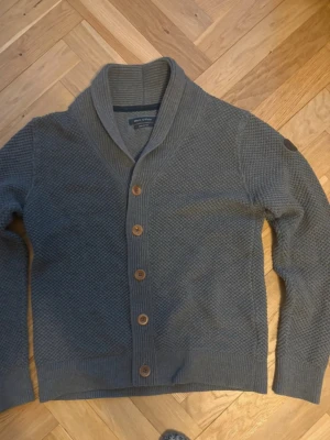 Grå Marco Polo kofta med sjalkrage och träknappar - Stilren grå kofta från Marco Polo med sjalkrage och träknappar framtill. Koftan har ett diskret strukturmönster, ribbade muddar och en patch på ena ärmen. Perfekt för lager på lager och en avslappnad look.