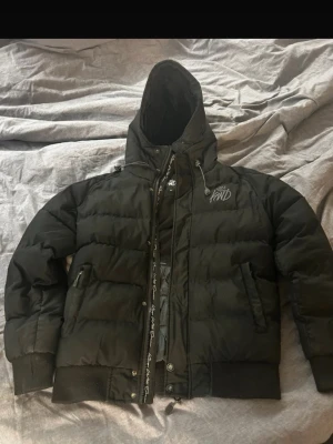Svart pufferjacka från KWD med huva - Säljer en svart pufferjacka från KWD med broderad logga på bröstet och huva. Jackan har dragkedja, knappar och ribbade muddar vid ärmslut och nederkant. Perfekt för kalla dagar och har praktiska fickor med dragkedja. Materialet är syntet och fyllningen är tjock och isolerande.