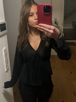 Svart plisserad blus med v-ringning - Snygg svart blus med plisserat tyg och v-ringning. Blusen har långa, vida ärmar och markerad midja som ger en feminin siluett. Perfekt att styla med både jeans och kostymbyxor. Materialet ser ut att vara ett glansigt syntetiskt tyg.