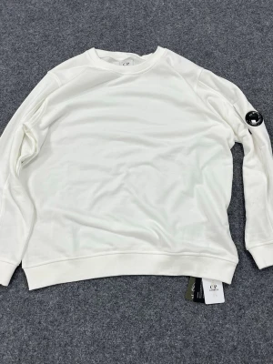 C.P Company Sweatshirt  - Snygg vit sweatshirt från C.P. Company med rund hals och ribbade muddar. Tröjan har en ikonisk svart linsdetalj på ena ärmen och är tillverkad i mjukt bomullsmaterial. Perfekt för en clean och stilren look.