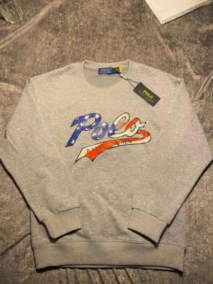 Grå sweatshirt från Polo Ralph Lauren - Snygg grå sweatshirt från Polo Ralph Lauren med stort tryck framtill i blått, vitt och rött. Klassisk rund halsringning och ribbade muddar vid ärmslut och nederkant. Perfekt för en avslappnad och trendig look. Skriv vid frågor och funderingar!