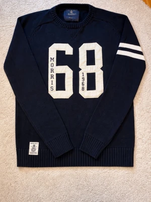Marinblå stickad tröja Morris - Snygg marinblå stickad tröja från Morris med stora vita siffror '68' och text på bröstet. Två vita ränder på ena ärmen ger en sportig känsla. Rund halsringning och ribbade muddar. Märkespatch nertill framtill. Perfekt för dig som gillar collegeinspirerad stil.