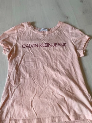 Ljusrosa Calvin Klein Jeans t-shirt - Säljer en ljusrosa t-shirt från Calvin Klein Jeans med tryckt logga i vinrött framtill. Klassisk rund halsringning och korta ärmar. Tillverkad i mjuk bomull som är skön att bära hela dagen.