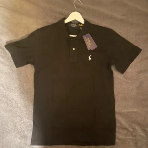 Svart pikétröja från Polo Ralph Lauren - Klassisk svart pikétröja från Polo Ralph Lauren med vit broderad logga på bröstet. Tröjan har korta ärmar, krage och två knappar framtill. Tillverkad i bomull och har en slim passform. Perfekt för en stilren och enkel look.