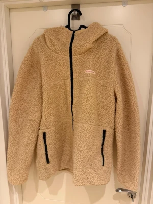 Beige teddyjacka Revolution - Mysig beige teddyjacka från Revolution med huva och dragkedja framtill. Jackan har två svarta dragkedjeförsedda fickor och Revolution-logga på bröstet. Tillverkad i fluffigt teddy-material som håller dig varm och bekväm.                                                                 Modellen är 185cm lång och väger 82kg.