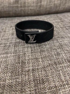 Louis Vuitton svart läderarmband - Snyggt svart armband från Louis Vuitton i läder med silverfärgat LV-spänne framtill. Armbandet har präglade monogramdetaljer längs bandet och ett stilrent metallspänne. Perfekt accessoar för dig som gillar exklusiv och tidlös design.