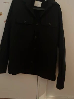 Svart overshirt med fickor (S) - Stilren svart overshirt med knäppning framtill och två bröstfickor med lock och knapp. Långärmad modell med klassisk krage, perfekt att slänga över en t-shirt. Från Jack and Jones (tror den har slutats säljas) Den är för stor för mig. Pris kan diskuteras vid snabb affär. Använd 1 gång 