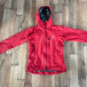 Röd vindjacka från Haglöfs med huva - Snygg röd vindjacka från Haglöfs med justerbar huva och dragkedja framtill. Jackan har två stora fickor med dragkedja och en bröstficka. Tillverkad i slitstarkt, vattenavvisande material och har reflexdetaljer på bröstet. Pris är ej hugget i sten