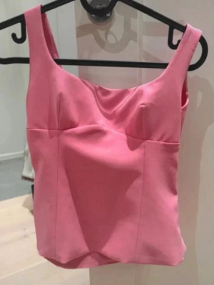 Rosa linnetopp med rosett i ryggen - Supersöt rosa linnetopp med breda axelband och en snygg rosettdetalj i ryggen. Axelbanden är uppsydda för att bli kortare som man ser på bild 4. Toppen har en figurnära passform och ärmlös design, perfekt för att styla med jeans eller kjol. 