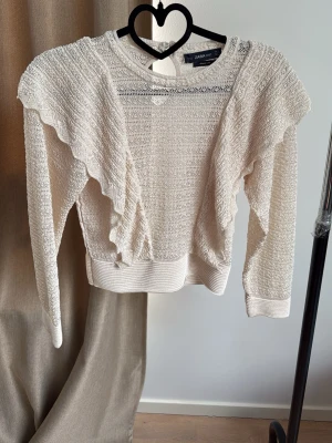 Beige stickad tröja med volanger Zara - Snygg beige stickad tröja från Zara med volangdetaljer framtill och längs ärmarna. Tröjan har rund halsringning, ribbade muddar och ett tunt, luftigt material som ger en trendig look.