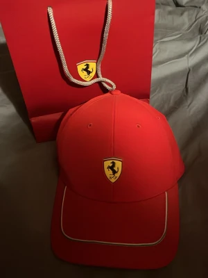 Ferrari keps - Snygg röd keps med Ferrari-logga framtill och Puma-logga på insidan. Klassisk böjd skärm med vit piping och justerbar passform. Perfekt för dig som gillar motorsport och vill sticka ut med en ikonisk accessoar. Köpt i Italien för två år sedan och har aldrig använts. Kvitto o påse följer med från Ferrari butiken i modena! 