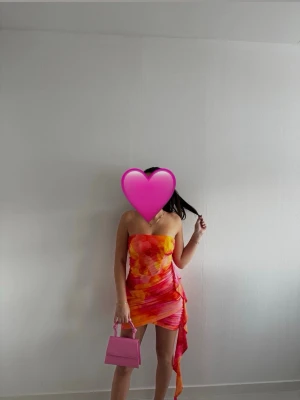 Färgstark axelbandslös kortklänning - Säljer en axelbandslös kortklänning i orange, rosa och gul med abstrakt mönster. Klänningen har omlottdetalj och volang på sidan, samt en figurnära passform. Perfekt för sommarens alla tillfällen!