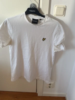 Vit t-shirt från Lyle & Scott - Klassisk vit t-shirt från Lyle & Scott med rund hals och det ikoniska gula örnlogot på bröstet. Tillverkad i mjuk bomull och har en enkel, clean design som passar till det mesta. Perfekt basplagg för alla tillfällen.